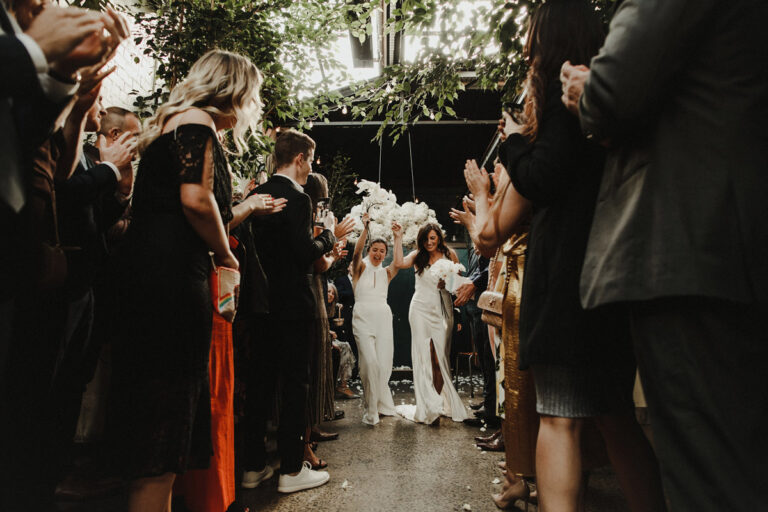 A joyous procession through a leafy corridor with guests clapping as two individuals walk down hand in hand.