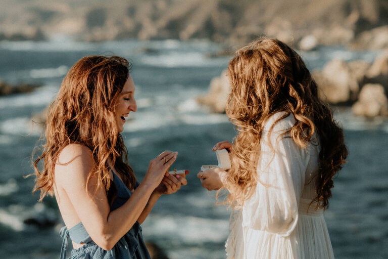 The couple smiles at each other with the ocean waves and cliffs creating a stunning backdrop.