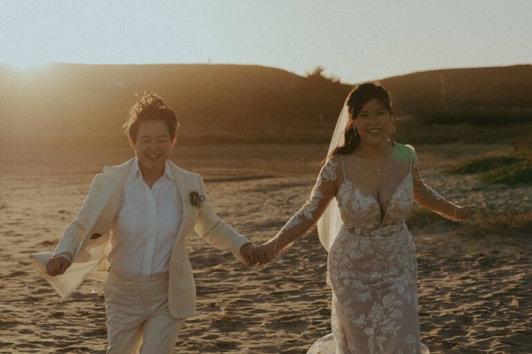 Two brides run barefoot across the sand at sunset after their Mavericks House Half Moon Bay wedding.