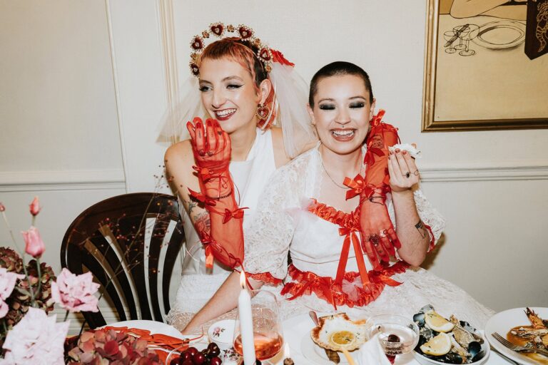A vibrant bouquet of red and blush flowers held against black weddingwear and a dark vintage wall.