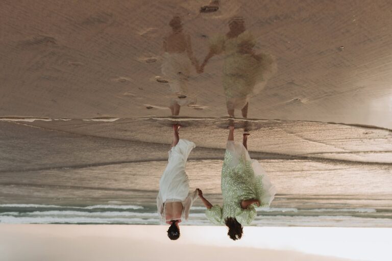 Two brides walk hand in hand across wet sand with their reflection clearly visible.