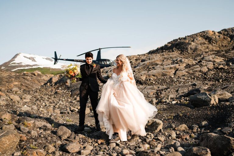 A couple walks over rocky terrain in front of a helicopter, wearing a floral gown and dark suit.