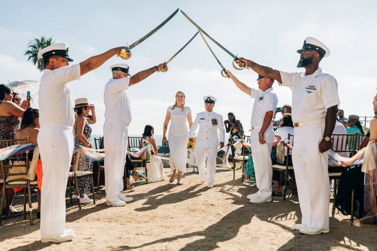 Queer couple walks hand-in-hand beneath raised military swords at their beach wedding ceremony exit.