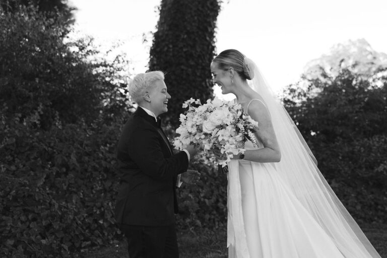 A bride and her partner smile at each other in a garden after their first look, holding a white bouquet.