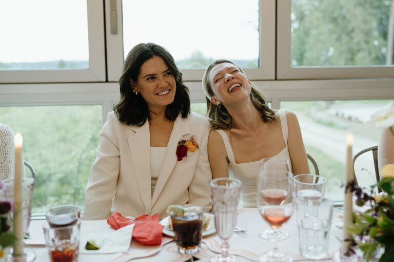 Two brides sit at their reception table, smiling and laughing with glasses and florals in front of them.