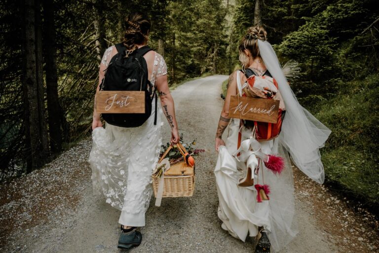Two queer brides walk down a forest path with backpacks, floral bouquet and picnic basket in hand.
