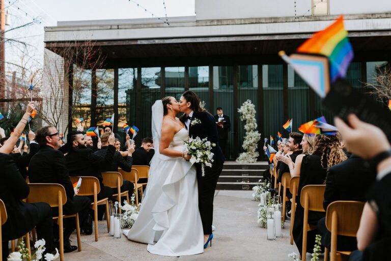 Couple kisses at end of wedding aisle while guests wave progress pride flags and cheer around them.