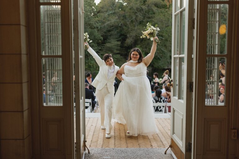 Two brides raise their bouquets and walk through arched doors just after their ceremony.