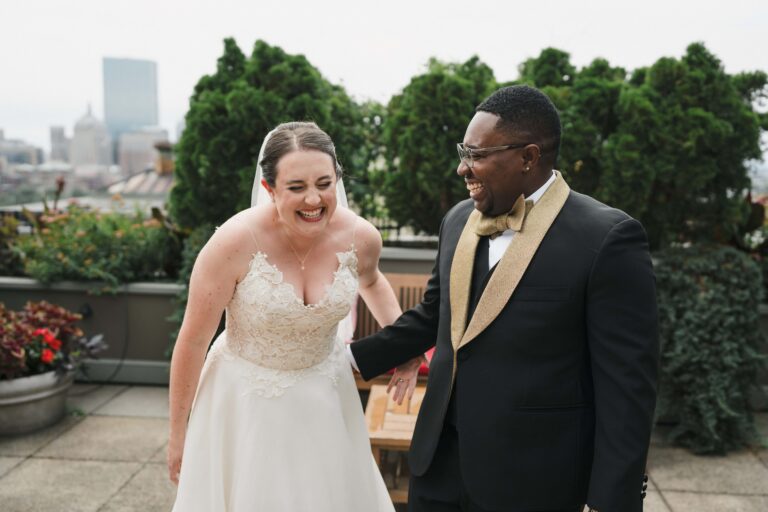Two people in wedding attire laugh together while standing on a rooftop patio with plants behind them.