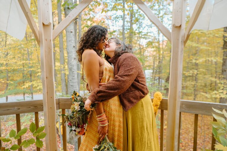 Two brides kiss beneath a wooden arch surrounded by autumn trees in Northern Michigan during their elopement.