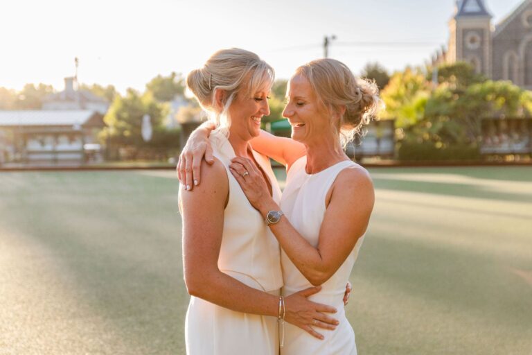Two brides embrace, smiling and holding each other closely on a lawn bowls green at sunset.