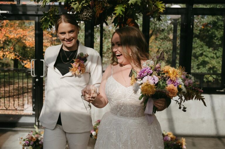 Two brides walk through their greenhouse ceremony space smiling and holding hands with bouquets