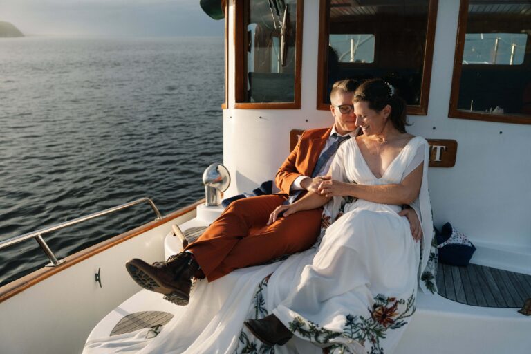 Two people in wedding outfits sit close on a white boat, surrounded by open water at sunset.