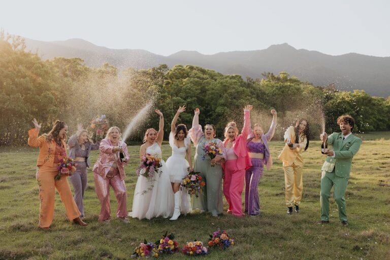 Wedding party dressed in bright suits celebrates with champagne on a Byron Bay field at sunset.