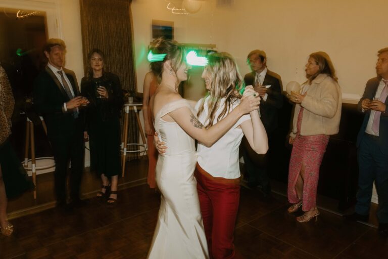 A couple shares their first dance, one in an ivory gown and one in red trousers and blouse.