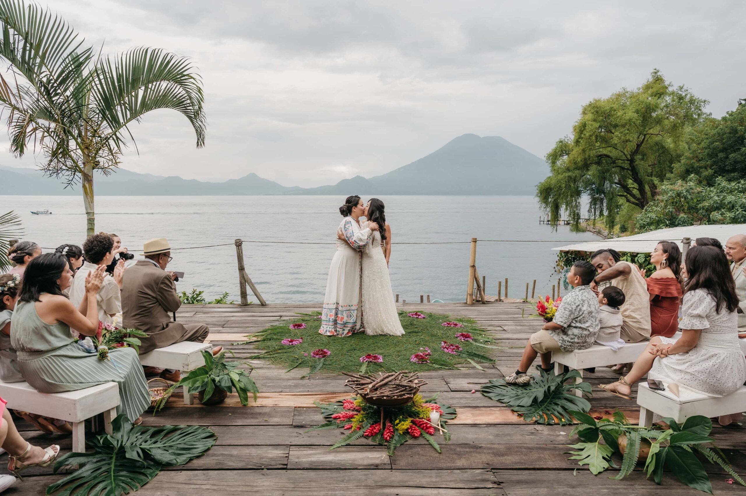 Newlywed brides kiss on petal ring altar overlooking Lake Atitlan while guests clap on wooden deck.