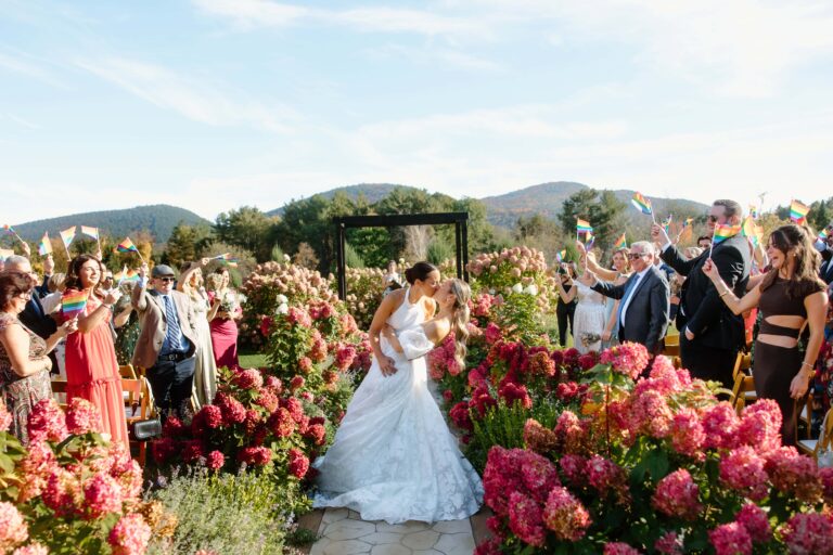 Newlyweds kiss on hydrangea lined aisle as guests wave rainbow flags against Adirondack mountain backdrop.