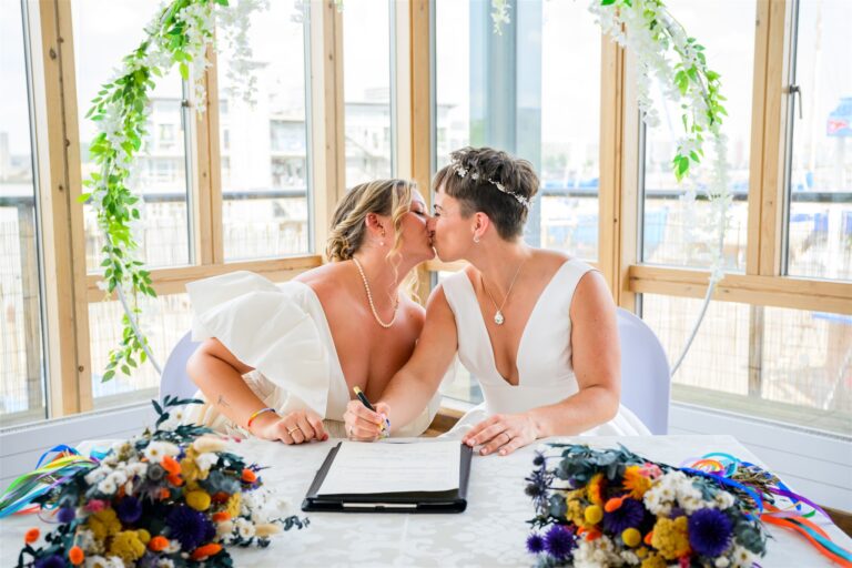 Newlyweds share kiss while signing registry, colourful dried bouquets and greenery hoop framing marina windows.