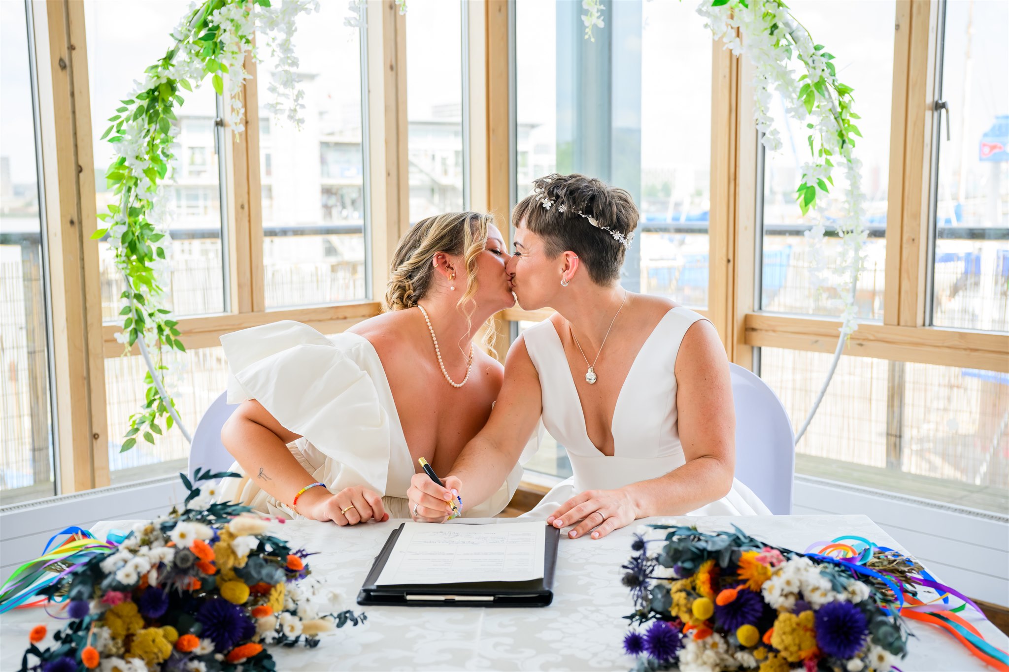 Newlyweds share kiss while signing registry, colourful dried bouquets and greenery hoop framing marina windows.