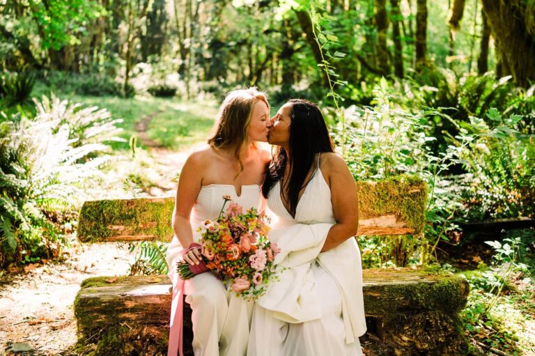 Two brides kiss on a rustic log bench covered in moss and surrounded by ferns.