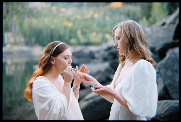 One person lights a joint for their partner as they stand in white dresses near a lake