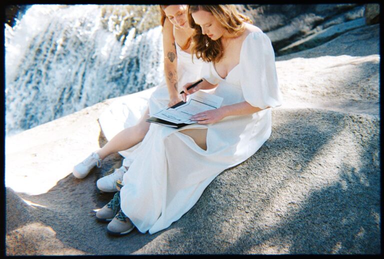 Two people in white dresses sit on a rock as one signs the marriage license near a waterfall