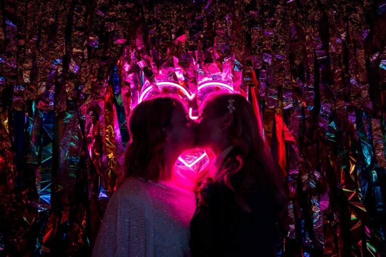 Bec and Lara kiss in front of a glowing pink neon heart with a foil streamer backdrop and rainbow lighting.