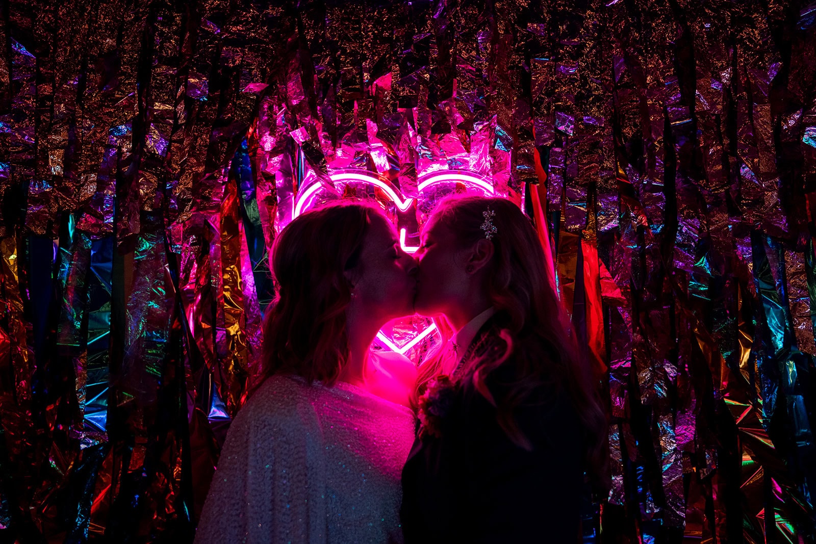 Bec and Lara kiss in front of a glowing pink neon heart with a foil streamer backdrop and rainbow lighting.