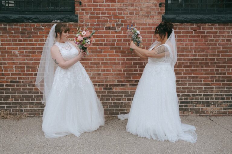 Iryelis and Sarah face each other playfully holding up their bouquets against a red brick wall.