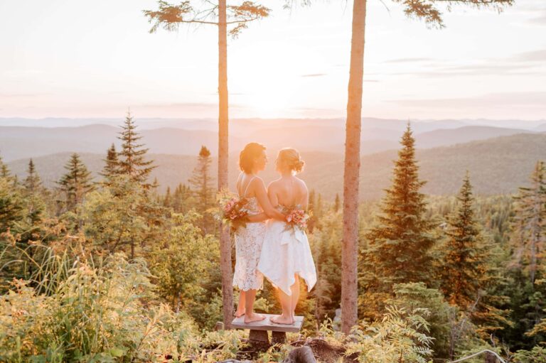 Two barefoot brides hold each other on a small platform, with sun setting over mountain landscape.