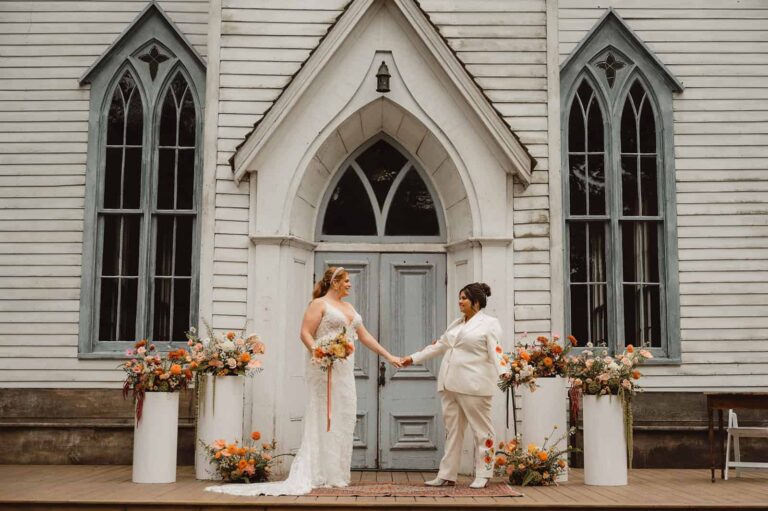 Two brides hold hands on a chapel porch framed by orange and cream floral columns and vintage architecture.