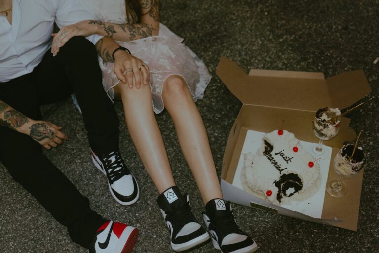 Couple sits on pavement in wedding outfits and Jordans beside smashed heart-shaped cake and desserts.