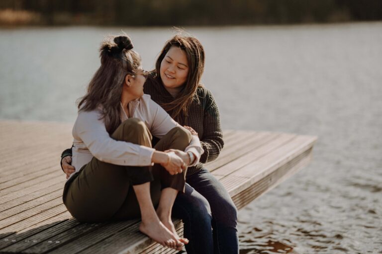 Two women sit close together on a wooden dock with water and trees in the background.