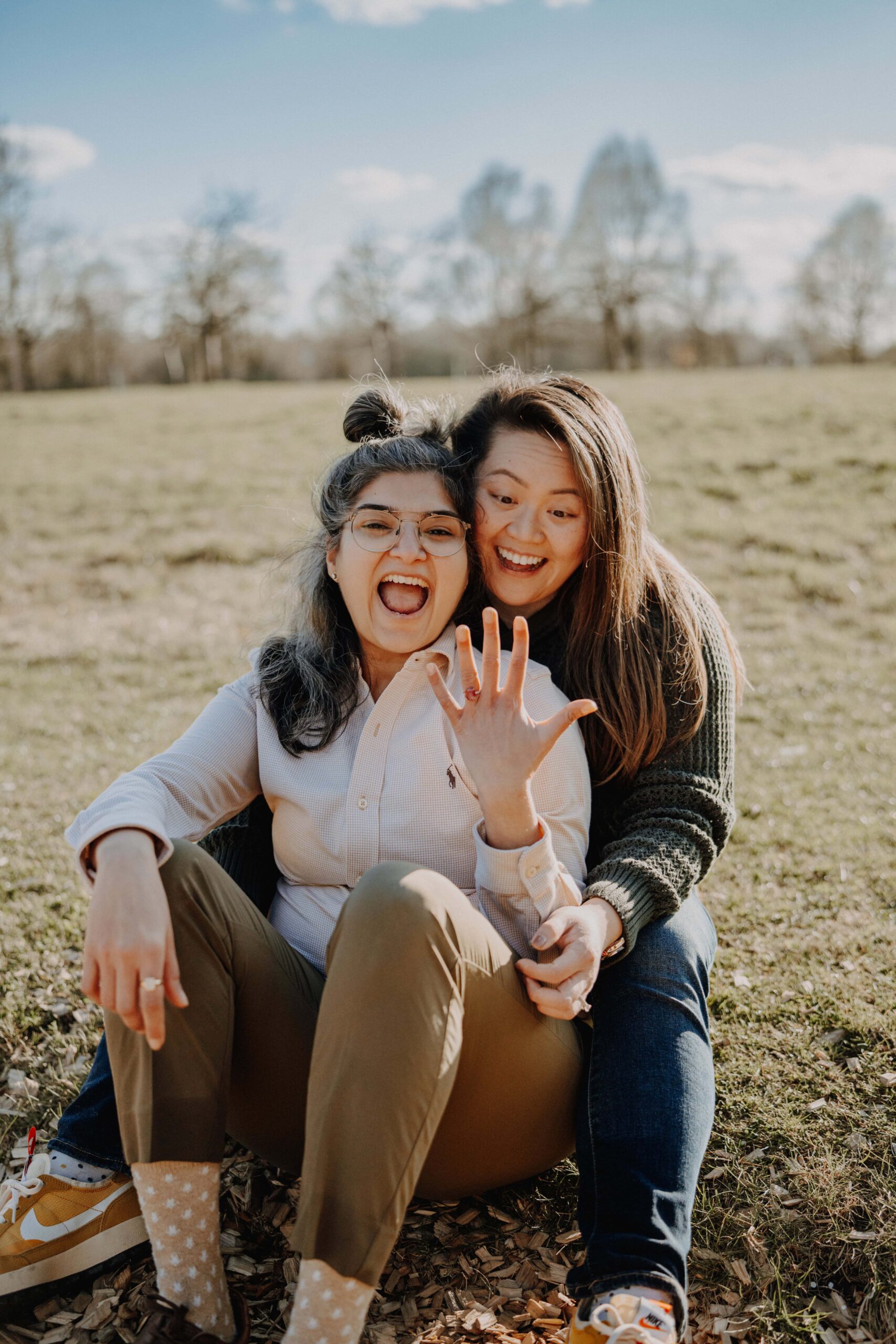 Two women sit together in a grassy park, one holding out her hand to show her ruby ring.