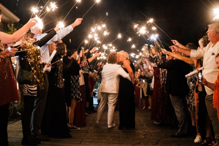 Two brides kiss mid-sparkler tunnel as guests cheer, waving gold pom-poms and sparklers.