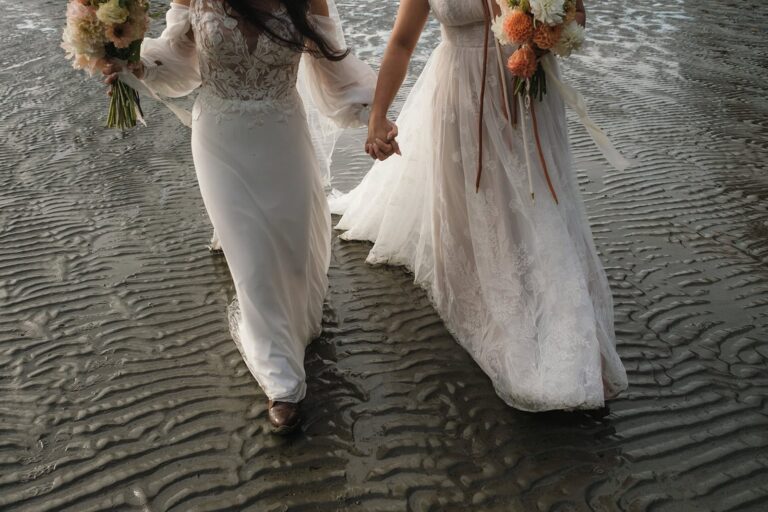 Two brides hold hands while walking across rippled wet sand, each holding a bouquet.