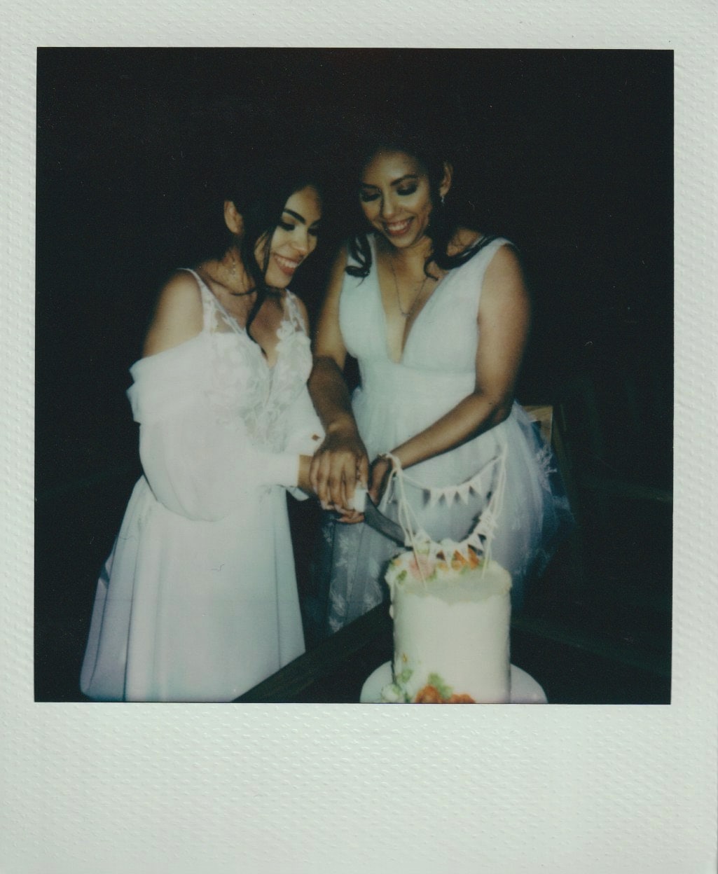 Two brides cut a single-tier white cake decorated with florals and a “just married” bunting topper.