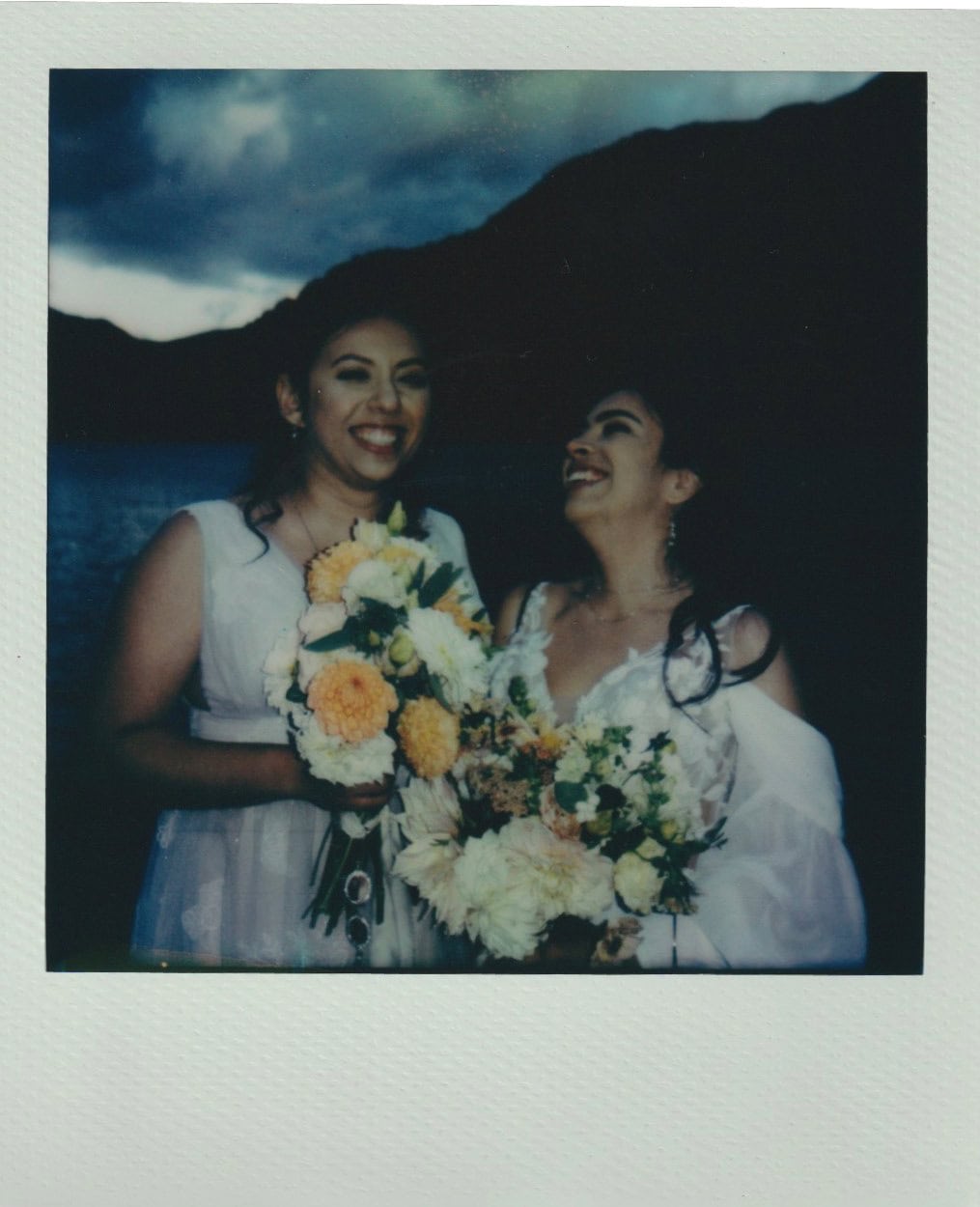 Two brides laugh together in front of a lake holding pastel-toned wedding bouquets.