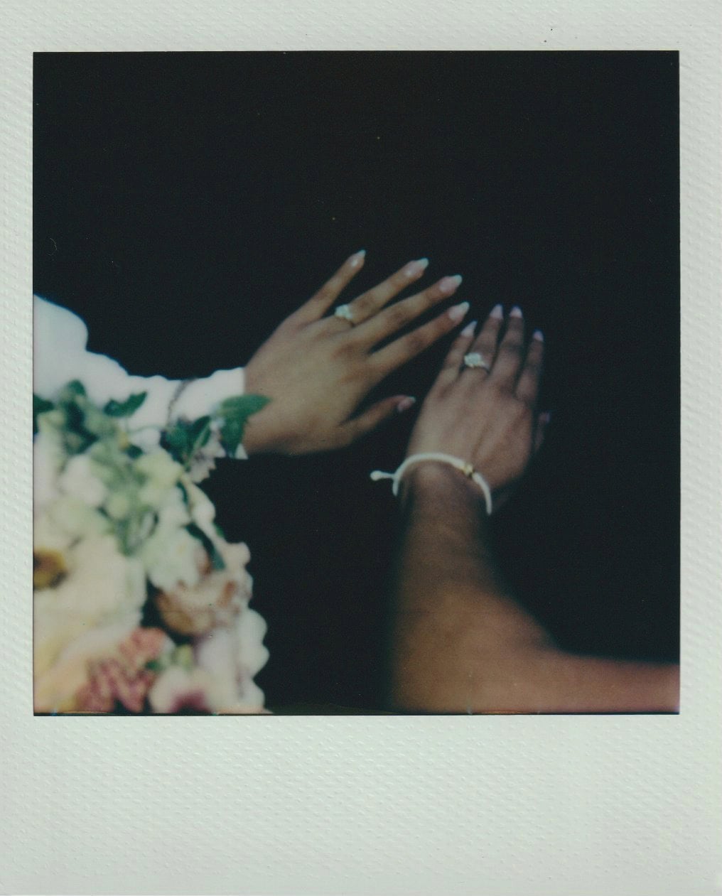 Two brides raise their hands to show wedding rings, with a bouquet of flowers partially in view.