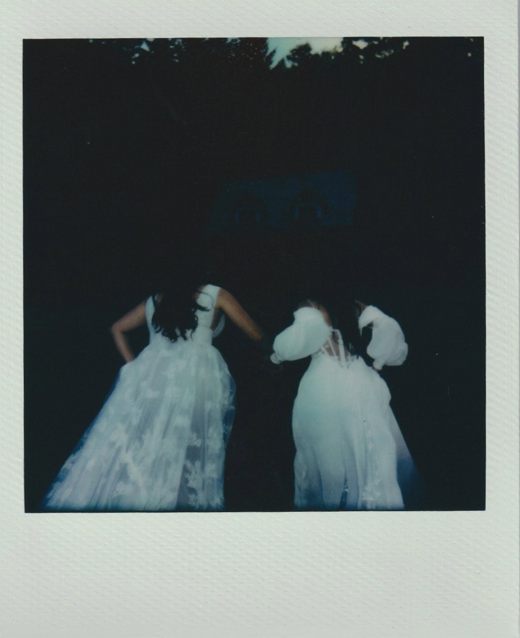 Two brides in white dresses walk together at dusk toward a log cabin surrounded by trees.