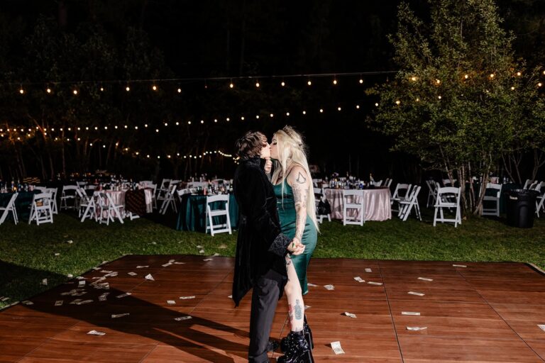Two brides kiss on a wooden dance floor under string lights, surrounded by scattered dollar bills.