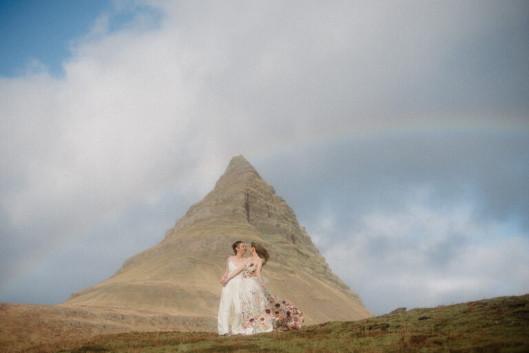 Two brides stand under a rainbow with a pointed Icelandic mountain rising behind them.