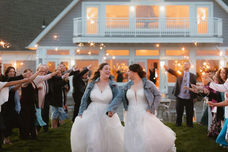 Brides wearing pearl-embellished denim jackets walk hand-in-hand through a sparkler send off outside the venue.