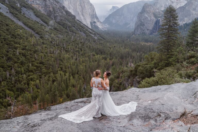 Two brides in lace wedding dresses stand on a rocky Yosemite cliff, overlooking pine-covered valleys and granite peaks.