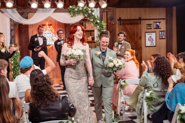 Two brides hold hands while walking down the aisle, guests clapping and floral décor framing the walkway.