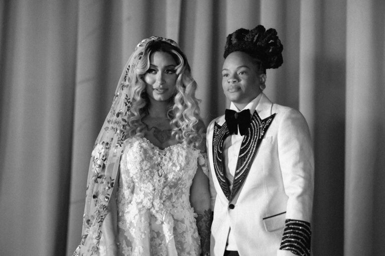 Bride wearing a floral embroidered veil stands next to her partner in a white beaded tuxedo jacket.