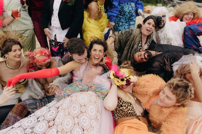 A joyful group lies together on the sand in colorful surrealist costumes at a Stradbroke Island wedding.