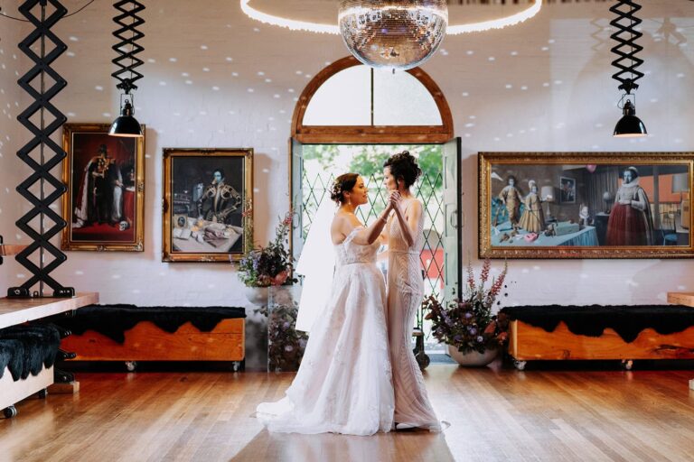 Two brides dance together under a disco ball in a venue with artwork-lined walls and wood floors.