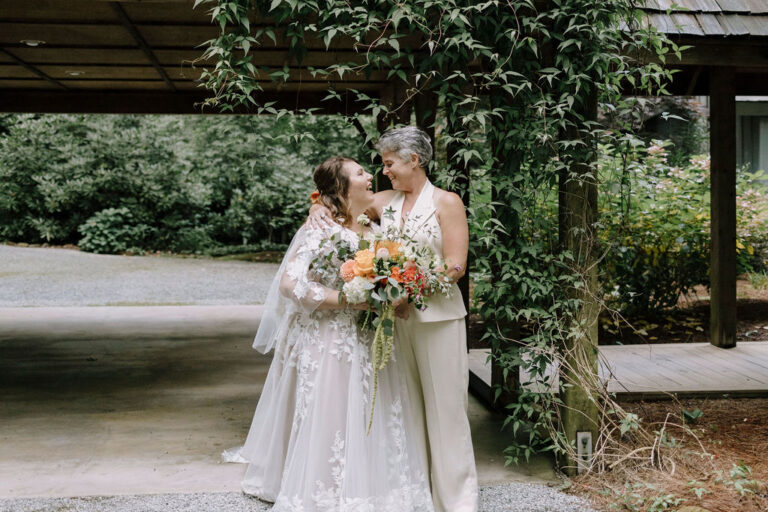 Couple stand under a wooden arbor, one holding a bouquet of orange, white, and red flowers.