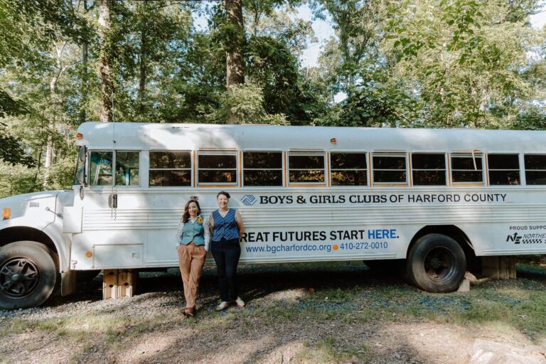 Two people stand leaning on a white Boys and Girls Club school bus parked in a wooded outdoor wedding venue.